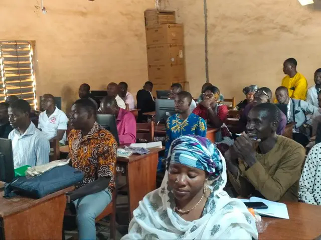 The image shows a group of people sitting at desks in a classroom, with monitors, bags, books,...