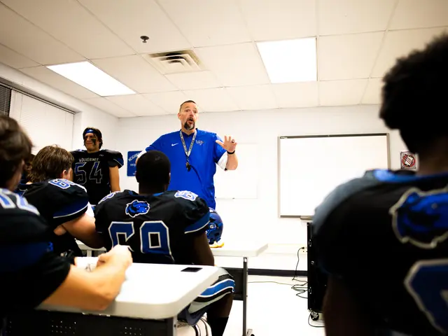 The image shows a man standing in front of a group of young men in a classroom. He is wearing a...