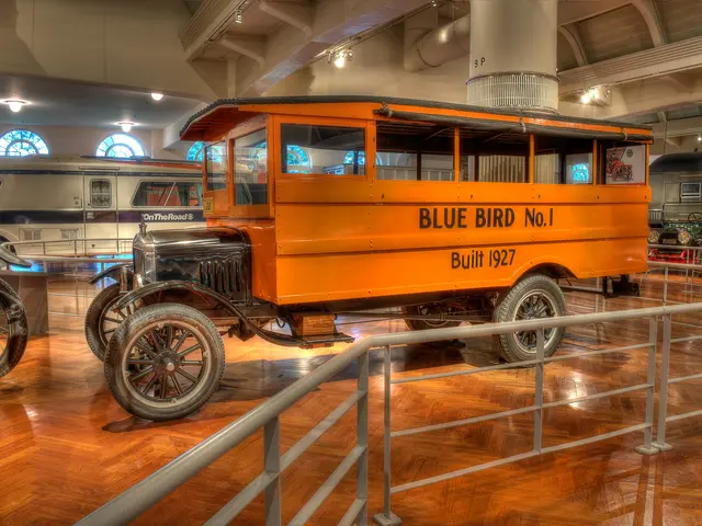 The image shows an orange bus on display in a museum, surrounded by other vehicles on the floor....