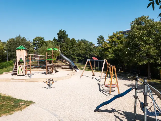 The image shows a playground with a slide and swings in the middle of it surrounded by grass,...