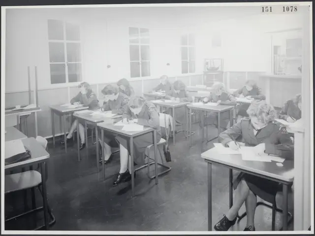 The image shows a black and white photo of a classroom full of students sitting at desks, writing...