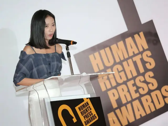 The image shows a woman standing at a podium in front of a microphone, with a banner behind her...
