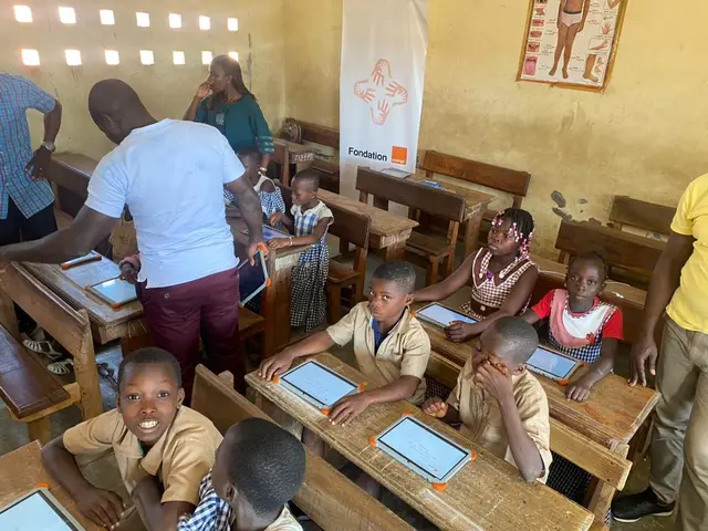 The image shows a group of children sitting at desks in a classroom, with a few people standing...