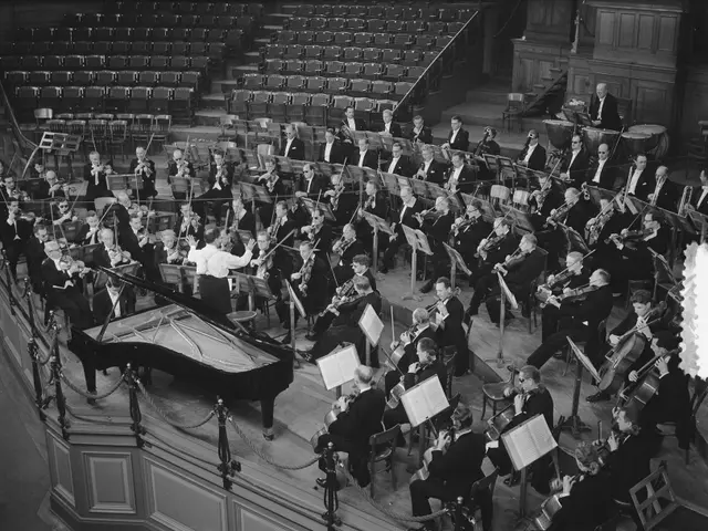The image shows a large orchestra in a large auditorium with a piano in the center. The orchestra...