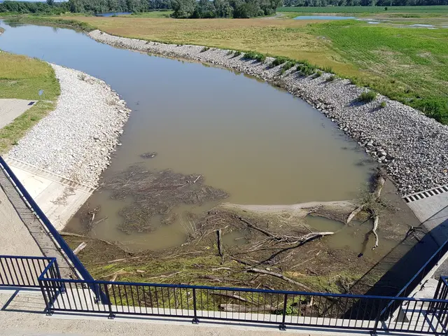 The image shows a river running through a lush green field next to a bridge, with railings, stones,...