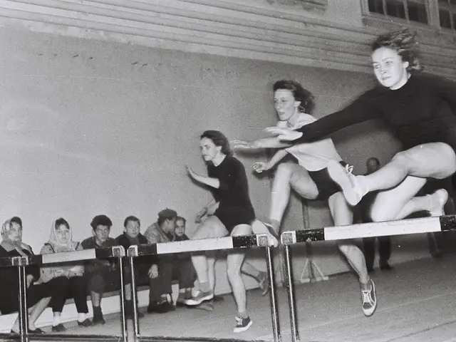 The image shows a black and white photo of two women jumping over a hurdle in a gymnasium. There...