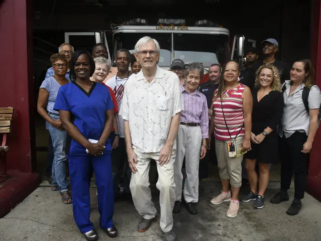 The image shows a group of people standing in front of a fire truck, with a bench on the left side...