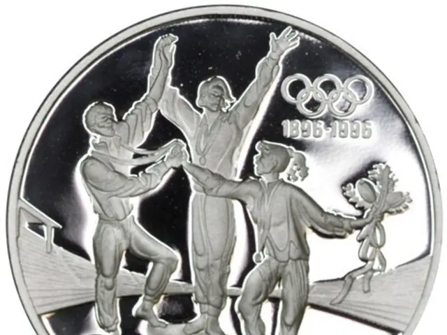 The image shows a silver medal with the Olympic logo on it, depicting three people standing on a...