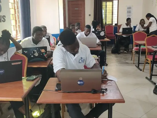 The image shows a group of people sitting at desks with laptops in a classroom. There are bags on...