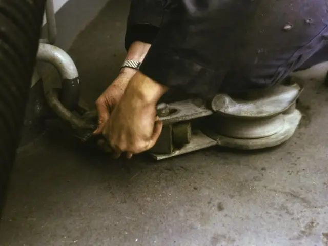 The image shows a man working on a machine in a garage, holding an object in his hands. He is...