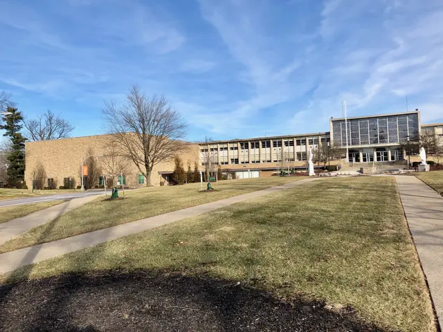 The image shows a large building with a walkway in front of it, surrounded by grass, trees, light...