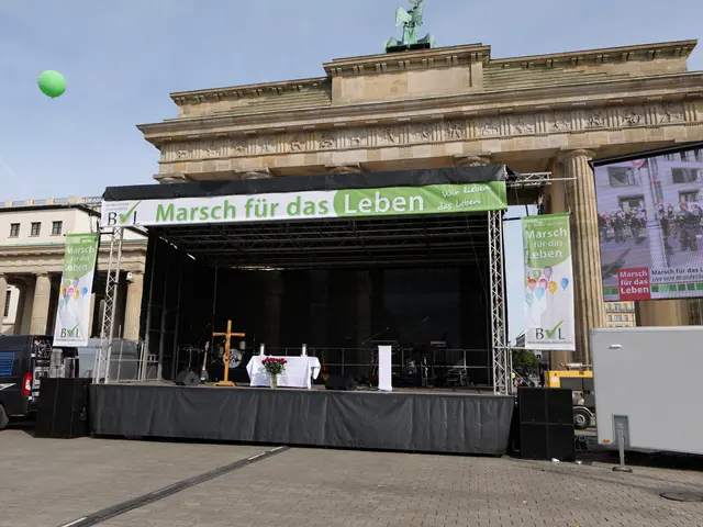 The image shows a stage set up in front of the Brandenburg Gate in Berlin, Germany. On the stage...