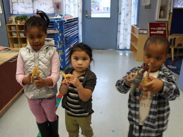 The image shows three children standing next to each other in a classroom, each holding a toy in...
