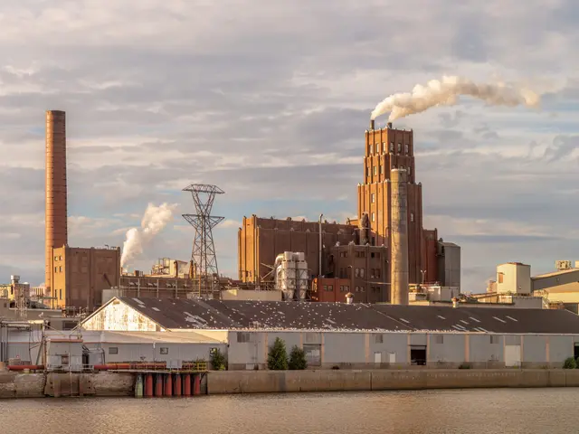 The image shows a large factory with smoke billowing out of its chimneys, situated next to a body...