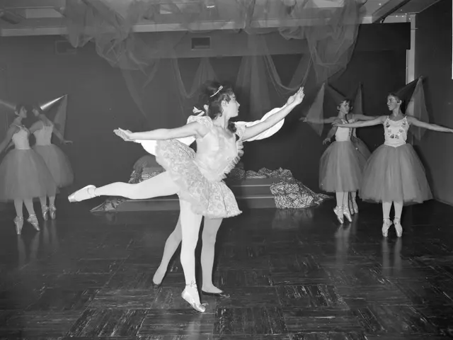 The image shows a group of young ballerinas in tutus and pointes dancing on a stage, with a wall in...