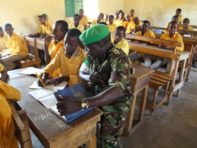 The image shows a group of children sitting at desks in a classroom, with books and pens on the...