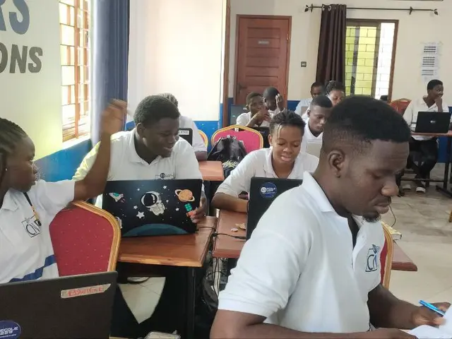 The image shows a group of people sitting at desks in a classroom, with laptops, books, bags, and...
