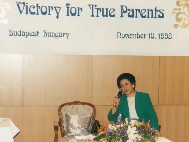 The image shows a woman sitting at a table with a microphone in her hand. On the table in front of...