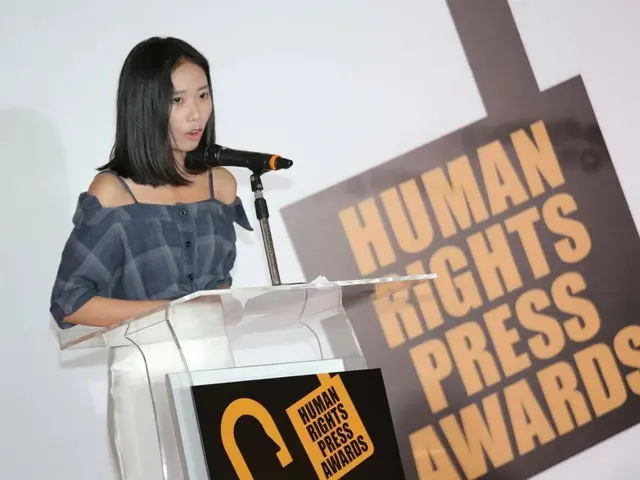 The image shows a woman standing at a podium in front of a microphone, with a banner behind her...