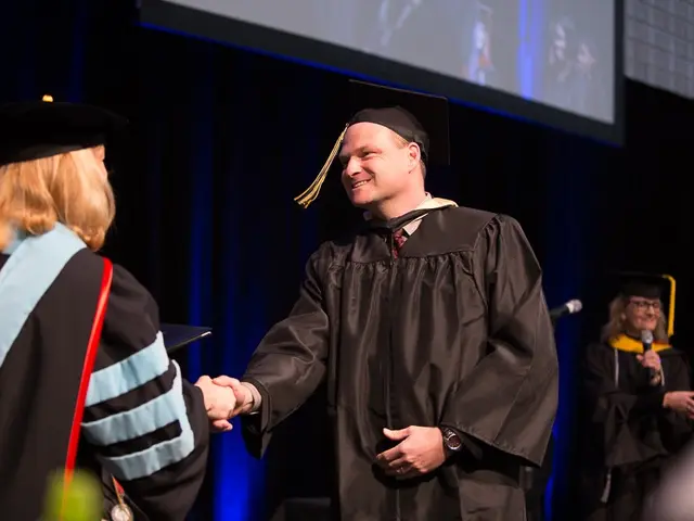 The image shows a man in a graduation cap and gown shaking hands with a woman in front of a podium,...