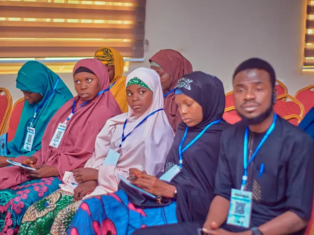 The image shows a group of people sitting in chairs in a room with a wall and window blinds in the...