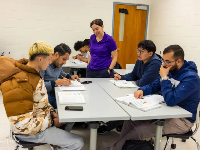 The image shows a group of people sitting around a table in a classroom. There are books, mobile...