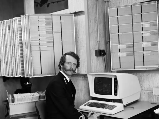 The image shows a man sitting at a desk in front of a computer, with a monitor, keyboard,...