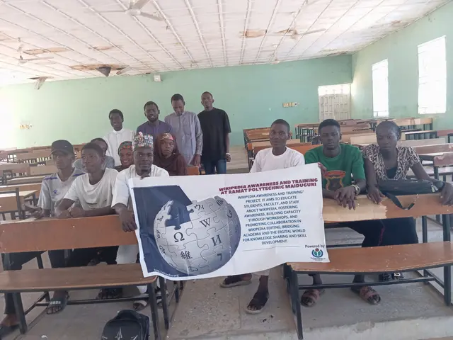 The image shows a group of people sitting at desks in a classroom, with a banner held up in front...