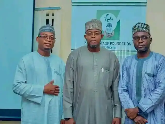 The image shows three men standing next to each other in front of a projector screen, wearing...
