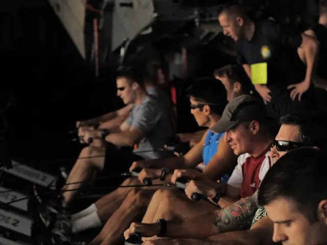 The image shows a group of people sitting on rowing machines in a gym, with a few people standing...