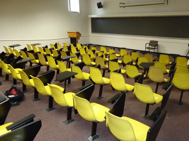 The image shows a lecture room with yellow chairs arranged in rows, tables, bags on the floor, a...