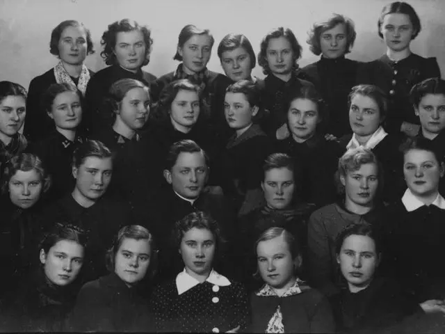 The image shows a black and white photo of a group of women in school uniforms, with a wall in the...