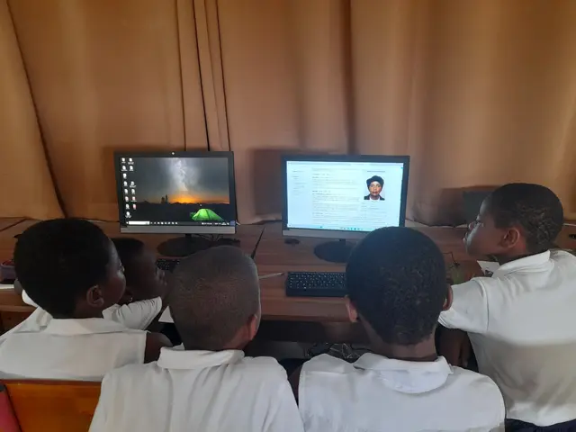 The image shows a group of children sitting at a desk in front of two computer monitors, with a...