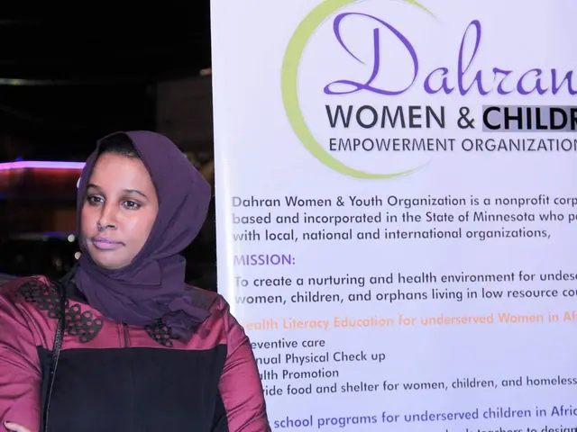 The image shows a woman standing in front of a banner that reads "Dahran Women & Children...