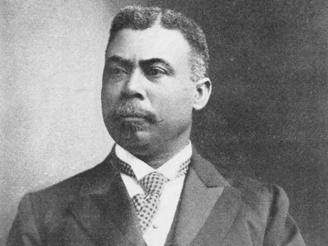 The image shows a black and white photo of a man wearing a blazer, shirt and tie, with a mustache,...