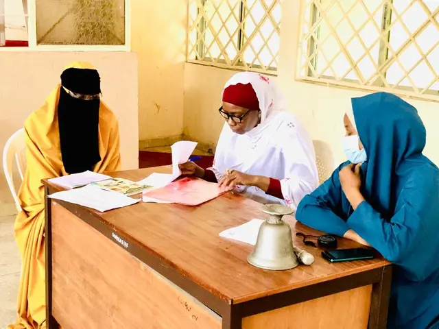 The image shows three women sitting at a table with papers, books, a bell, a mobile phone, and...