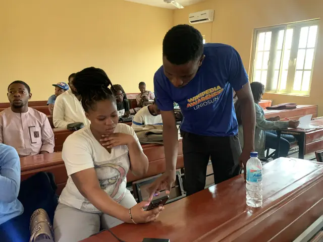 The image shows a group of people sitting at desks in a classroom, with a man standing and holding...