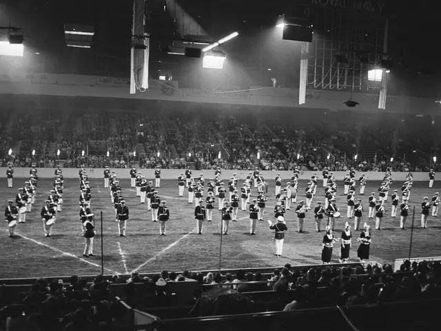The image shows a black and white photo of a marching band performing in a stadium. There are many...