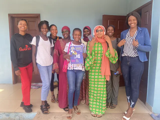 The image shows a group of women standing next to each other in front of a building, smiling and...