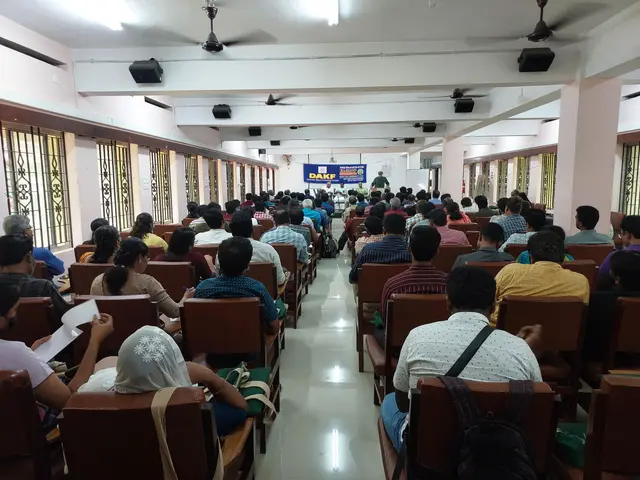 The image shows a large group of people sitting in chairs in a room with windows on the left and...
