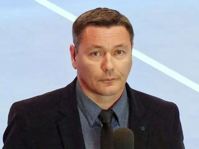 The image shows a man in a suit and tie holding a microphone, standing in front of a screen. He has...