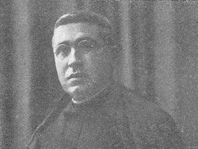 The image shows a black and white photo of a man in a priest's robes, wearing glasses and standing...