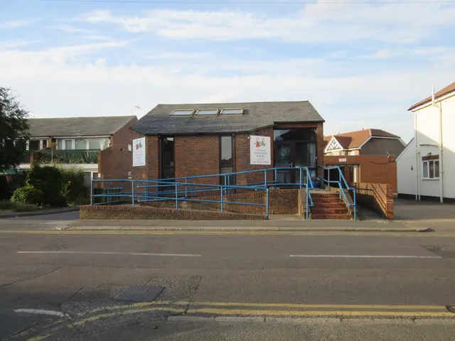 The image shows a school building with a blue railing in front of it, surrounded by houses with...