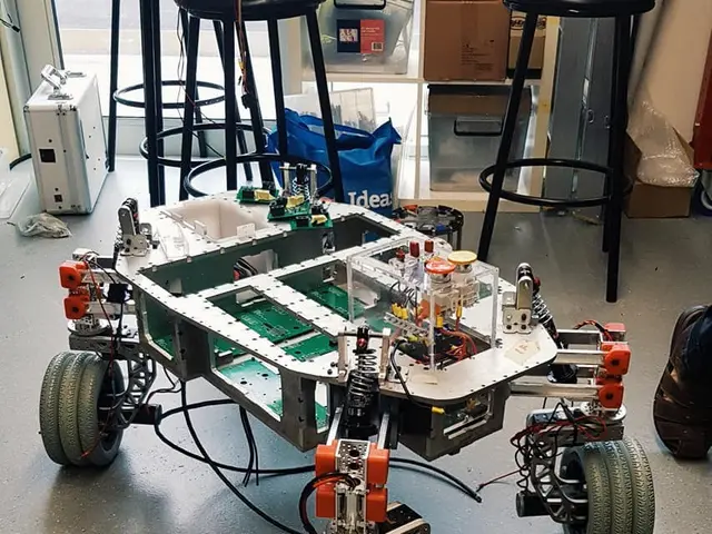 The image shows a robot sitting on top of a table in a room, surrounded by stools, cardboard boxes,...