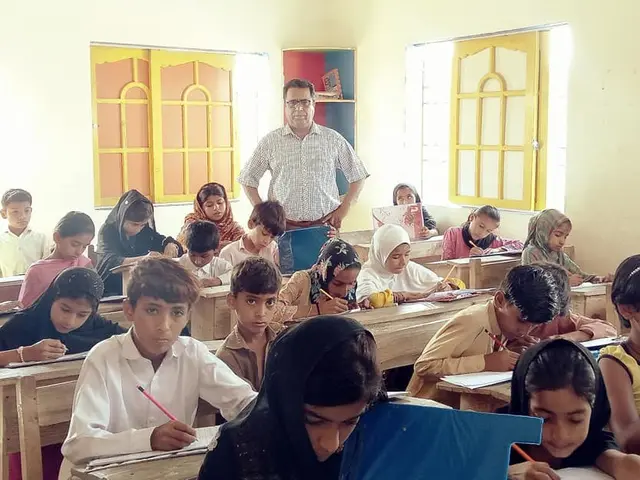The image shows a group of children sitting at desks in a classroom, with books and pens in their...