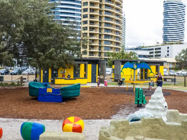 The image shows a playground in the middle of a city with tall buildings in the background. There...