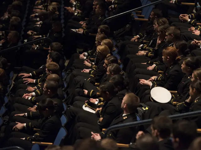 The image shows a large group of people in military uniforms sitting in an auditorium, with...