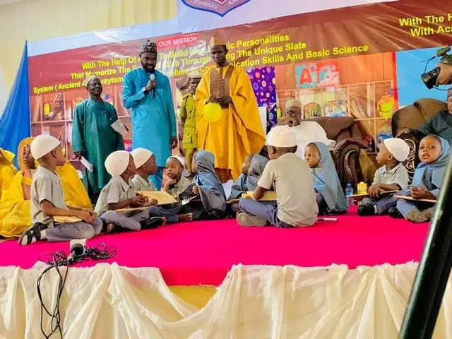 The image shows a group of children sitting on top of a stage, with some of them holding books in...