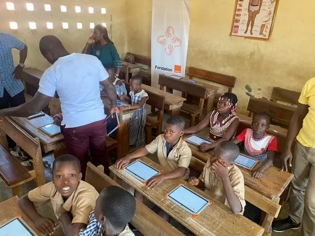 The image shows a group of children sitting at desks in a classroom, with a few people standing...