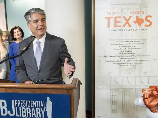 The image shows a man wearing a blazer, tie, and shirt standing at a podium in front of a banner...